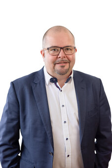 Geschäftsführer Tobias Lang. Person mit Brille, Kinnbart und kurz geschorenem Haar trägt blauen Anzug und weißes Hemd, schaut frontal in die Kamera; neutrales Porträt vor weißem Hintergrund, Oberkörperansicht.