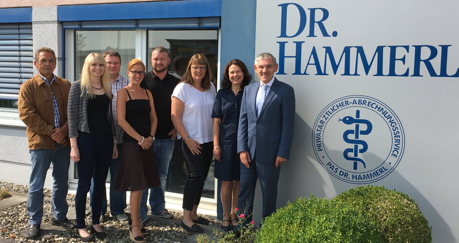 Gruppe von acht Erwachsenen steht nebeneinander und blickt in die Kamera vor Gebäude mit Aufschrift: „DR. HAMMERL“. Rundlogo daneben: „PRIVATÄRZTLICHER-ABRECHNUNGSSERVICE · P.A.S. DR. HAMMERL“. Fenster, Eingangstür, Kiesbeet und Büsche.