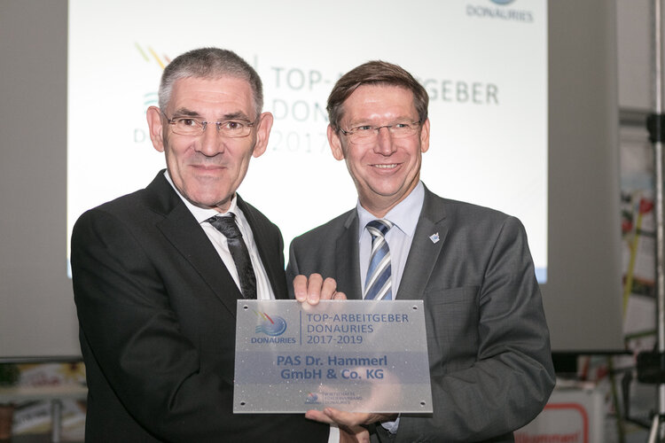 PAS Geschäftsführer Dr. Siegfried Hammer und Landrat Stefan Rössle in Anzügen halten eine Auszeichnungsplakette vor einer Projektionsleinwand. Auf der Plakette steht: "TOP-ARBEITGEBER DONAURIES 2017–2019 – PAS Dr. Hammerl GmbH & Co. KG".