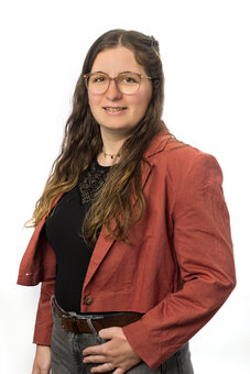 Person mit langen, welligen Haaren und Brille steht frontal und blickt in die Kamera; eine Hand am Gürtel. Studio-Porträt vor weißem Hintergrund, rostroter Blazer, schwarzes Oberteil mit Spitze, graue Hose.