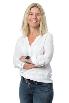 Person mit schulterlangem blondem Haar steht mit verschränkten Armen und lächelt in die Kamera; trägt weiße Bluse, blaue Jeans und Smartwatch, vor neutralem weißem Hintergrund, Porträtaufnahme.