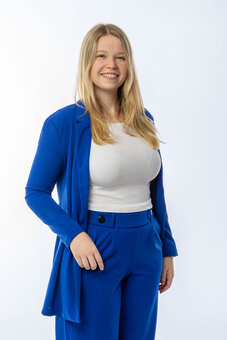 Frau steht frontal und lächelt in die Kamera, hält mit einer Hand ihren offenen blauen Blazer. Trägt weißes Oberteil und blaue Anzughose vor hellem, neutralem Studiohintergrund.