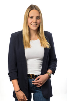 Person mit langen blonden Haaren steht frontal und lächelt in die Kamera; trägt dunkelblauen Blazer, weißes Top, blaue Jeans, schwarzen Gürtel mit goldener Doppelschnalle; neutraler, weißer Studiohintergrund.