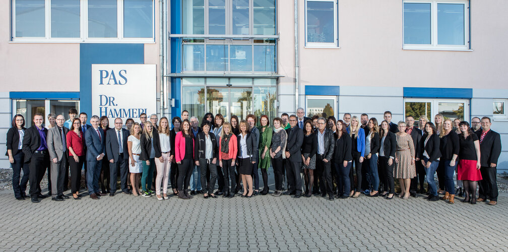 Große Gruppe Erwachsener steht nebeneinander und blickt zur Kamera, posierend für ein Gruppenfoto vor einem Bürogebäude mit Glaseingang; links ein Schild mit Text: “PAS” und “DR. HAMMEL”.