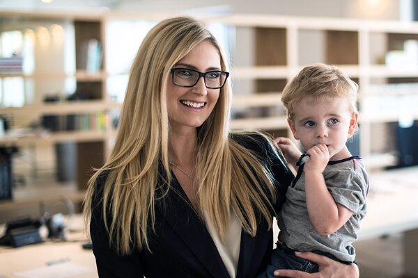 Erwachsene Person mit langen blonden Haaren und Brille hält ein kleines Kind auf dem Arm; das Kind nuckelt am Finger. Helles, modernes Büro mit Regalen und Schreibtischen im unscharfen Hintergrund.