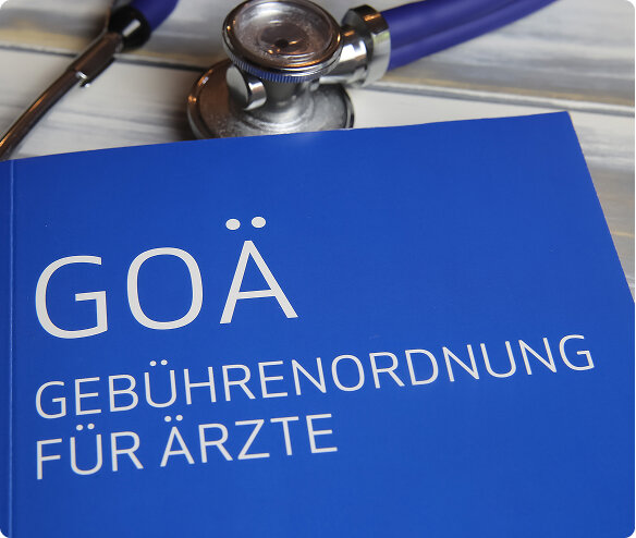 Blaues Heft mit Aufdruck GOÄ GEBÜHRENORDNUNG FÜR ÄRZTE liegt auf heller Holzoberfläche; daneben ein Stethoskop mit metallischem Kopf und blau-violettem Schlauch, Nahaufnahme.