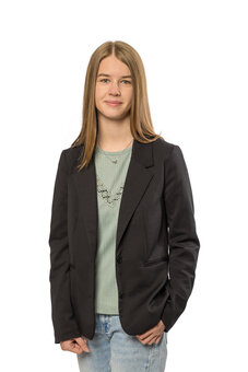 Person mit schulterlangem, glattem Haar trägt dunkelblauen Blazer über hellgrünem T‑Shirt und Jeans, steht frontal und blickt in die Kamera; Studioaufnahme vor weißem Hintergrund.