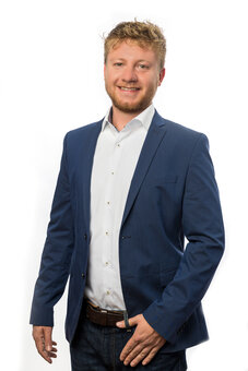 Mann im blauen Sakko, weißem Hemd und dunkler Jeans steht frontal, lächelt und hält eine Hand in der Hosentasche. Studio-Porträt vor weißem Hintergrund, bis Oberschenkel sichtbar.