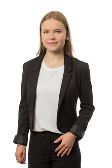 Person im schwarzen Blazer und weißem T‑Shirt steht frontal, blickt neutral in die Kamera und berührt mit der rechten Hand die Jacke; Halbporträt, hellem Hintergrund, Studioaufnahme.