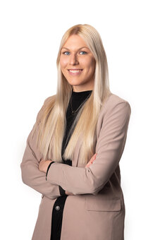 Person mit langen, glatten, blonden Haaren steht frontal mit verschränkten Armen, lächelnd. Trägt beigefarbenen Blazer über schwarzem Oberteil und Halskette. Studio-Porträt vor weißem Hintergrund, Oberkörperaufnahme.
