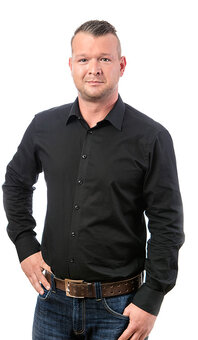 Mann in schwarzem Hemd steht mit einer Hand an der Hüfte und blickt in die Kamera. Studioaufnahme vor weißem Hintergrund, blaue Jeans, brauner Ledergürtel, Dreiviertelporträt.