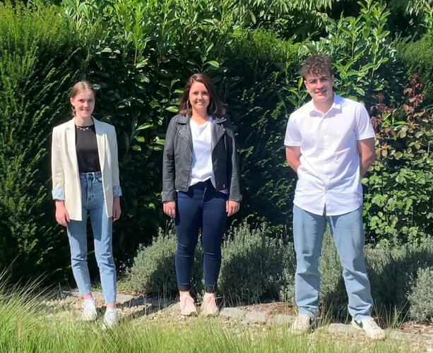 Drei Personen stehen nebeneinander und schauen in die Kamera, im Freien vor dichter grüner Hecke, auf kiesigem Boden, bei Sonnenschein, lässig gekleidet in Jeans, Jacken und weißen Sneakers.