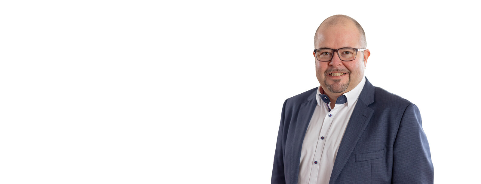 Geschäftsführer Tobias Lang mit Brille, kurzem Bart, im blauen Anzug und weißem Hemd lächelt frontal in die Kamera; Oberkörperporträt rechts im Bild, viel weißer Leerraum links, neutraler Studiohintergrund.
