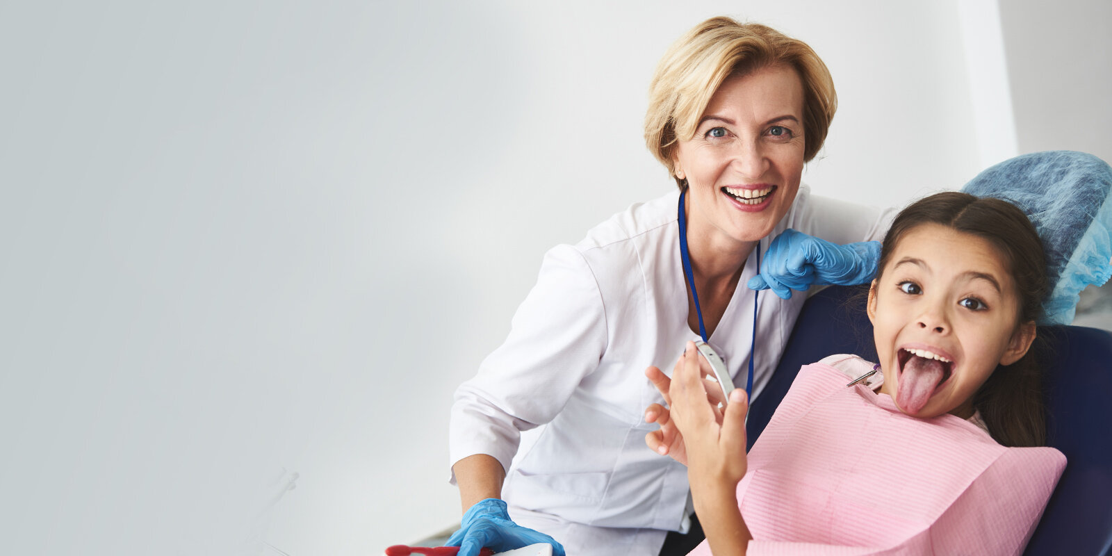 Zahnärztin mit blauen Handschuhen beugt sich über ein Kind im Behandlungsstuhl; das Kind trägt rosa Latz, hält einen Speichelsauger und streckt die Zunge heraus; helle Praxisumgebung.