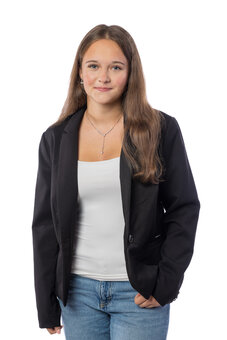 Person im Halbporträt steht frontal, eine Hand in der Hosentasche, Blick zur Kamera. Trägt schwarzen Blazer, weißes Top, blaue Jeans, Halskette. Neutraler Studiohintergrund, gleichmäßige Ausleuchtung.