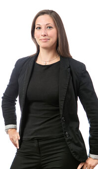 Stehende erwachsene Person in schwarzer Businesskleidung blickt in die Kamera, eine Hand auf der Hüfte. Langes braunes Haar. Studioaufnahme vor weißem Hintergrund, neutral ausgeleuchtet, Halbporträt.