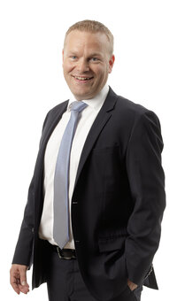 Mann im dunklen Anzug steht leicht seitlich und lächelt in die Kamera, eine Hand in der Hosentasche; helles Hemd und hellblaue Krawatte vor weißem Studiohintergrund.