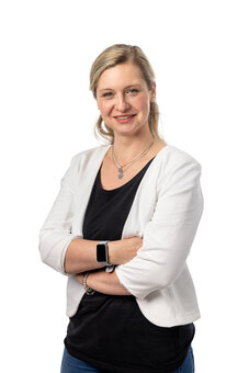 Erwachsene Person mit hellblondem Haar steht mit verschränkten Armen und blickt in die Kamera; weißer Blazer über schwarzem Top, Halsketten und Smartwatch; Studioaufnahme vor weißem Hintergrund.