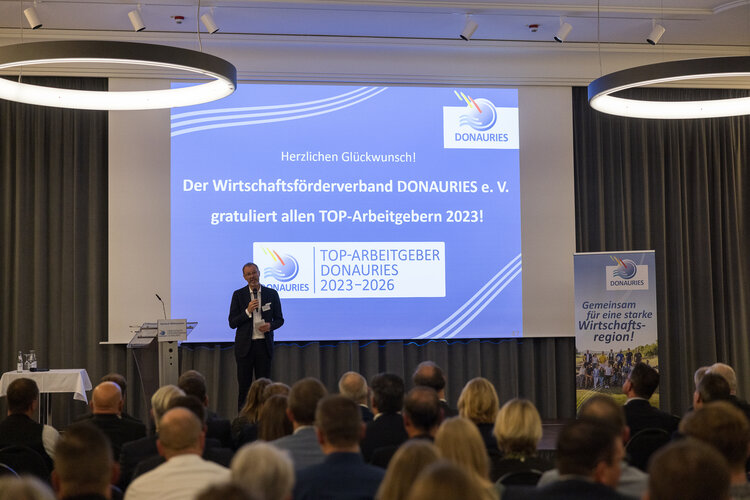 Präsentationsleinwand zeigt: „DONAURIES“, „Herzlichen Glückwunsch! Der Wirtschaftsförderverband DONAURIES e. V. gratuliert allen TOP-Arbeitgebern 2023!“, „TOP-ARBEITGEBER DONAURIES 2023–2026“. Redner mit Mikrofon präsentiert am Pult; sitzendes Publikum im Konferenzsaal; rechts Roll-up-Banner mit „DONAURIES“ und „Gemeinsam für eine starke Wirtschaftsregion!“.