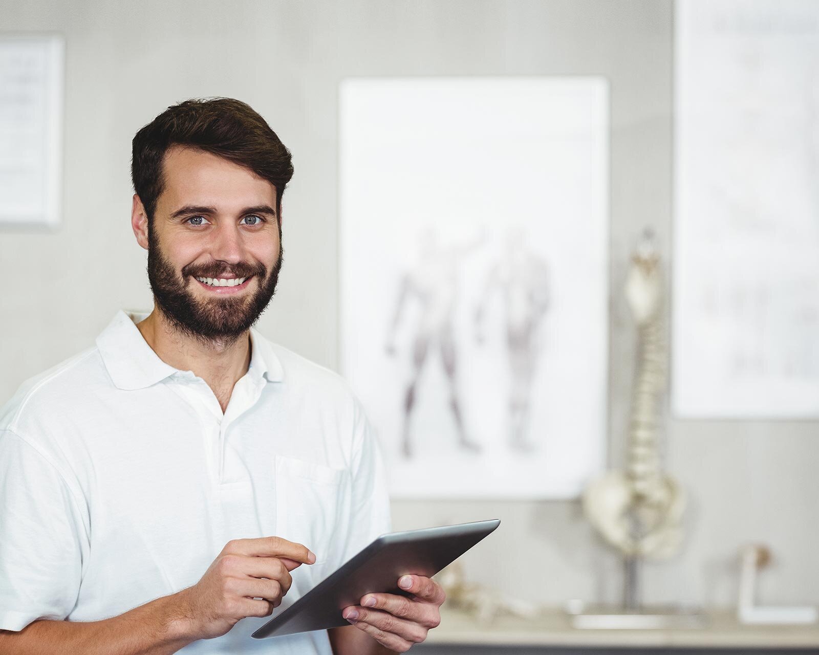 Bärtiger Mann im weißen Polohemd hält und bedient ein Tablet, blickt in die Kamera; im Hintergrund unscharfe anatomische Poster und ein Wirbelsäulenmodell in einem hellen Raum.
