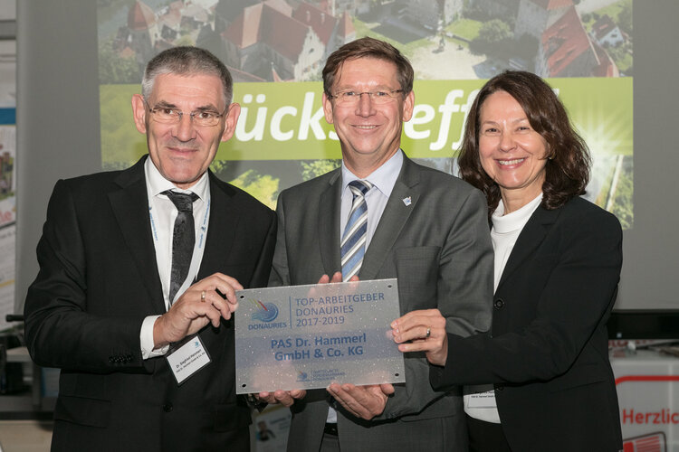 PAS Geschäftsführer Dr. Siegfried Hammerl links, Landrat Stefan Rössle mittig und Margarete Hammerl in Businesskleidung halten eine rechteckige Metallplakette; Innenraum mit Poster und Luftbild im Hintergrund. Text auf der Plakette: DONAURIES; TOP-ARBEITGEBER DONAURIES 2017-2019; PAS Dr. Hammerl GmbH & Co. KG.