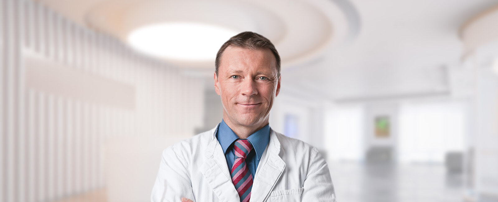 Dr. Mark Tanner in weißem Kittel mit Krawatte steht frontal mit verschränkten Armen und blickt in die Kamera; unscharfer, heller Innenraum mit runder Deckenleuchte, großen Fenstern und Flur.