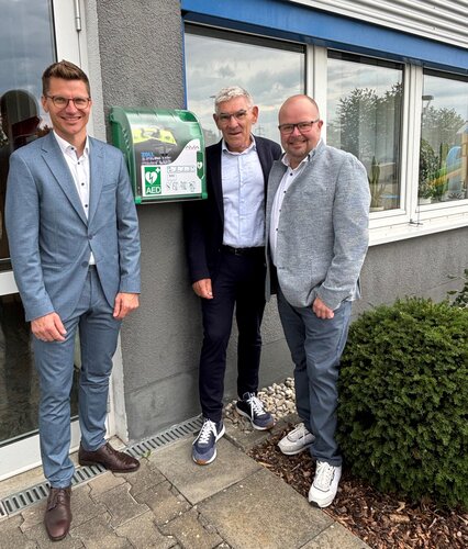 Grüner Defibrillator-Schrank mit Aufdruck "AED" und Herz-Blitz-Symbol hängt an einer Außenwand, während drei Männer in Anzügen daneben stehen; Umgebung mit Fensterfront, Eingangstür, Pflaster, Kies und Busch.