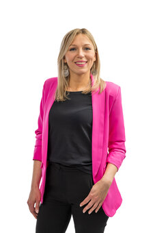 Erwachsene Person im pinken Blazer steht frontal und lächelt in die Kamera; trägt schwarzes T‑Shirt, schwarze Hose und silberne Ohrringe vor reinweißem Hintergrund, Hände locker an den Seiten.