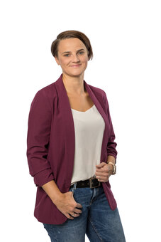 Stehende erwachsene Person mit kurzen braunen Haaren blickt in die Kamera; eine Hand in der Jeanstasche, die andere am Gürtel. Bordeauxfarbener Blazer, weißes Top, schwarze Uhr, Studiohintergrund weiß.