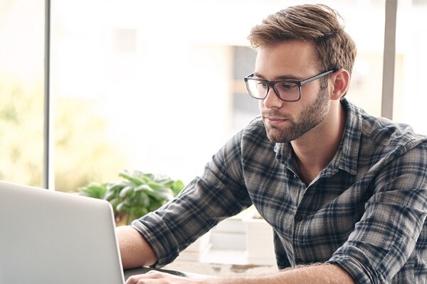 Person mit Brille arbeitet am Laptop, blickt auf den Bildschirm und tippt. Heller Innenraum mit Schreibtisch, Topfpflanze und großem Fenster, weiches Tageslicht im Hintergrund.