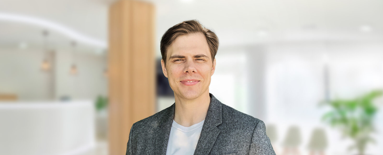 Alexander Frauenhoff mit kurzem braunem Haar im grauen Blazer und weißem T‑Shirt steht frontal zur Kamera; unscharfe, helle Bürolobby mit weißem Empfangstresen, Hängelampen, Holzsäule, Pflanzen und Stühlen im Hintergrund.