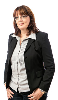 Person mit schulterlangem braunem Haar und Brille steht frontal und blickt in die Kamera, eine Hand in der Hosentasche. Trägt schwarzen Blazer, hellgraue Bluse, Jeans; weißer Studiohintergrund.