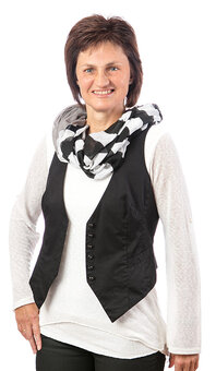 Person steht frontal und lächelt in die Kamera, trägt weißes Langarmshirt, schwarze Weste, schwarz-weiß gemusterten Schal und dunkle Hose; neutraler weißer Hintergrund, Arme locker seitlich.