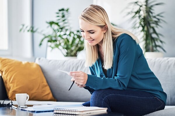 Frau sitzt auf einem Sofa, hält Zettel und Stift und betrachtet das Papier. Auf dem Couchtisch liegen Notizbuch, Unterlagen und Kaffeetasse; helles Wohnzimmer mit Pflanzen und Kissen.