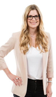 Person mit langen blonden Haaren und schwarzer Brille steht halbseitig, lächelt und stützt eine Hand in die Hüfte; beigefarbener Blazer, weißes Oberteil, dunkle Hose, neutraler weißer Hintergrund.