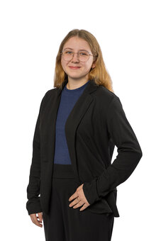 Person mit schulterlangem blonden Haar, Brille, trägt schwarzen Blazer, dunkelblaues Oberteil und schwarze Hose; steht frontal leicht gedreht, rechte Hand an der Jacke; weißer Studiohintergrund.