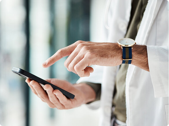 Person in weißem Laborkittel hält ein Smartphone und tippt mit dem Zeigefinger darauf. Nahaufnahme der Hände; Armbanduhr mit blauem Band; heller, unscharfer Innenraum mit Glasflächen im Hintergrund.