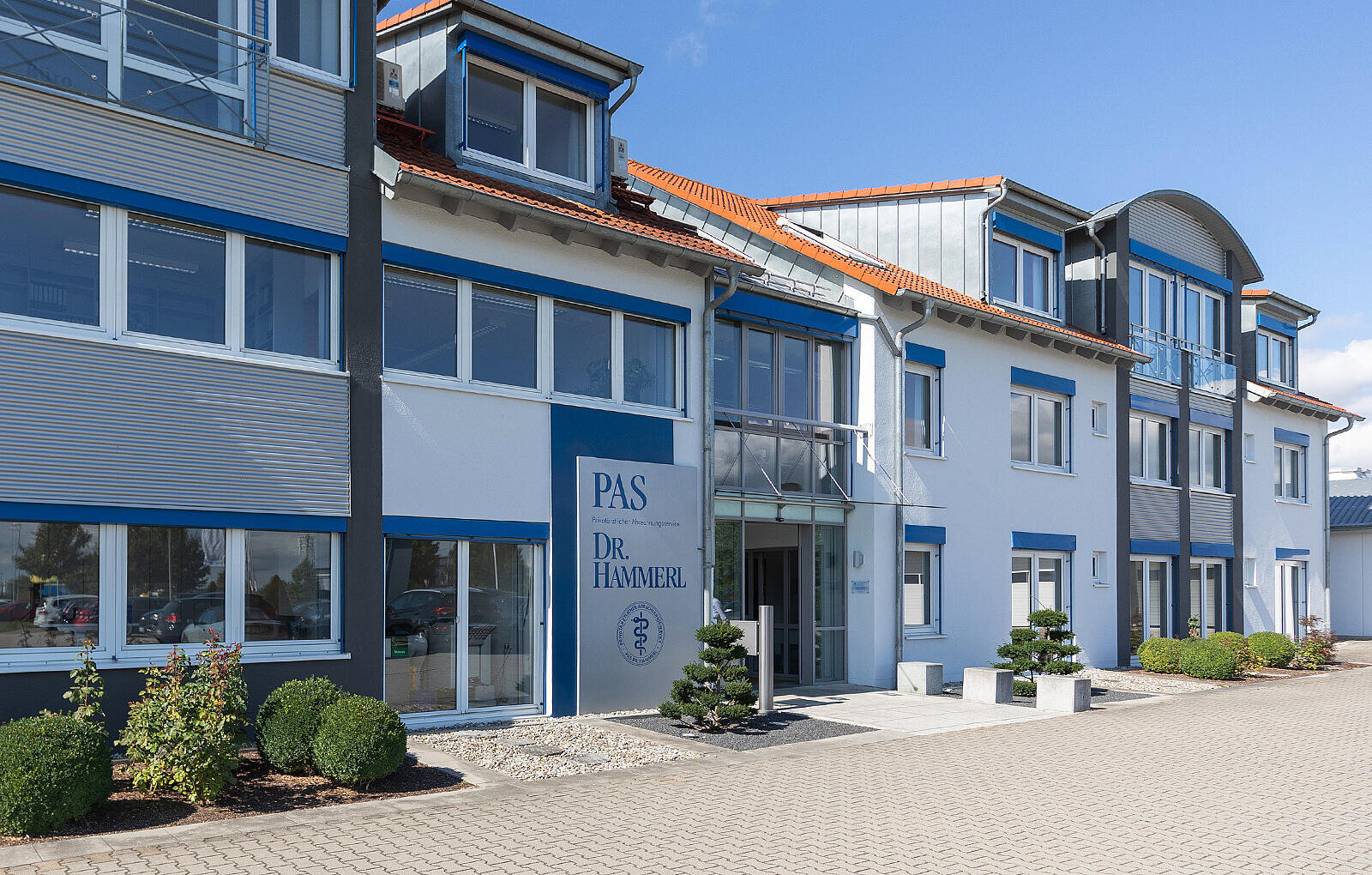 PAS Firmengebäude mit verglastem Haupteingang zeigt großes Schild mit Text: PAS und DR. HAMMERL; umgeben von weiß-blauer Fassade, roten Dachziegeln, Ziersträuchern und gepflastertem Vorplatz unter blauem Himmel.