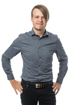 Mann steht frontal mit Händen in die Hüften gestützt, blickt in die Kamera. Graues langärmliges Hemd, schwarze Hose mit Gürtel. Studioaufnahme vor weißem Hintergrund, Oberkörper bis Oberschenkel sichtbar.