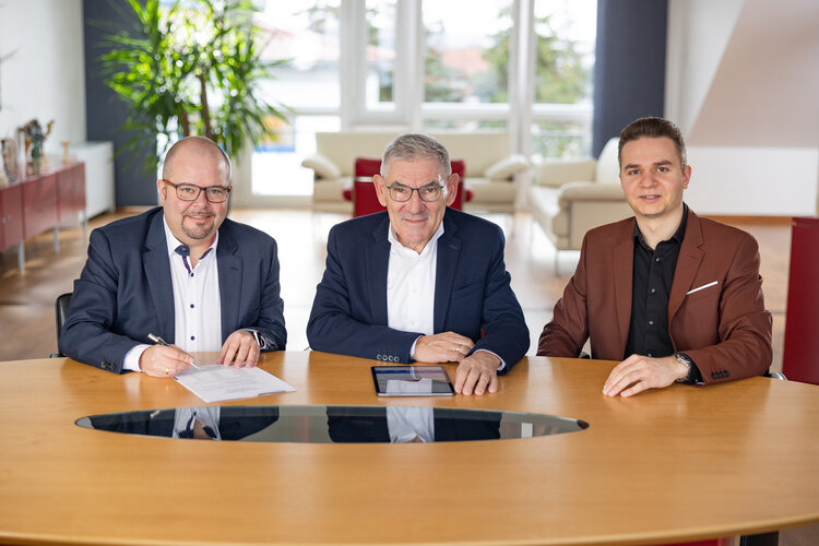 Drei Personen in Businesskleidung sitzen an einem Konferenztisch und blicken in die Kamera; links hält jemand einen Stift über ein Dokument, mittig liegt ein Tablet. Büro mit Fenstern, Pflanzen, Sofas.