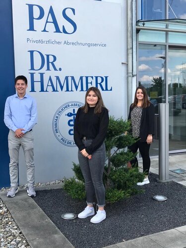 Drei Personen stehen nebeneinander vor einem Firmengebäude und einem großen Schild mit der Aufschrift: PAS Privatärztlicher Abrechnungsservice DR. HAMMERL; Glasfassade, kleiner Zierbaum und Kiesstreifen im Vordergrund.