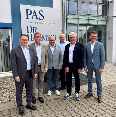 Sechs Männer stehen nebeneinander und blicken in die Kamera, vor einem Gebäudeeingang mit Glasfassade, Pflastersteinen und einem Firmenschild: "PAS Privatärztlicher Abrechnungsservice DR HAMMERL".