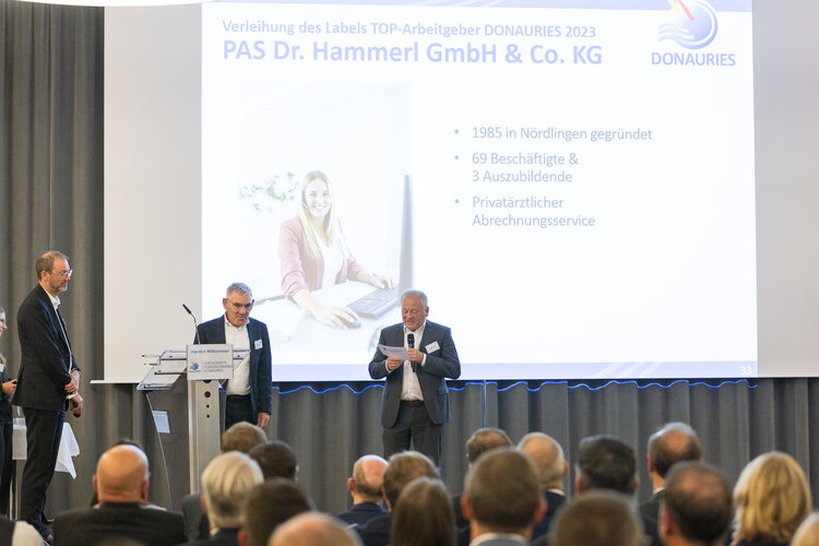 Bühne mit drei Personen vor Projektionsleinwand. Sie halten eine Ansprache; eine Person liest vom Blatt. Publikum sitzt; neben einem Foto einer Frau am Schreibtisch steht: Verleihung des Labels TOP-Arbeitgeber DONAURIES 2023; PAS Dr. Hammerl GmbH & Co. KG; DONAURIES; 1985 in Nördlingen gegründet; 69 Beschäftigte & 3 Auszubildende; Privatärztlicher Abrechnungsservice; 33.