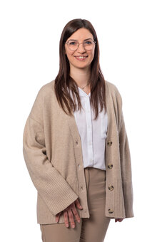 Erwachsene Person mit langen braunen Haaren und Brille steht frontal, blickt in die Kamera; trägt beigefarbenen Cardigan mit Knopfleiste, weiße Bluse und beige Hose; freigestellter weißer Hintergrund.