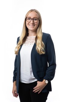 Person mit langen blonden Haaren und Brille steht und blickt in die Kamera; trägt dunkelblauen Blazer, weiße Bluse, schwarze Hose, Armbanduhr; eine Hand in der Hosentasche; Studioaufnahme vor weißem Hintergrund.