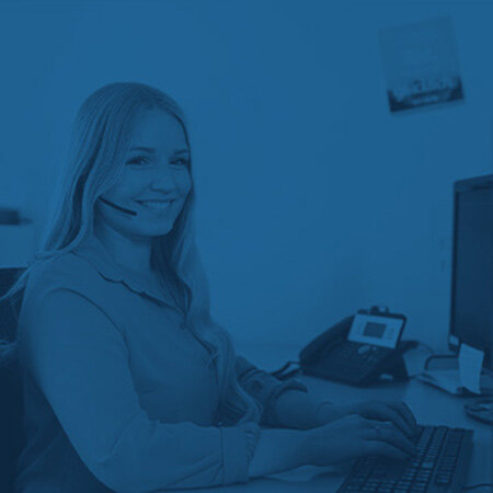 Person mit Headset sitzt am Schreibtisch, tippt auf einer Tastatur und blickt nach vorn. Im Bürohintergrund stehen Monitor, Tischtelefon, Maus, Notizzettel; das Bild ist monochrom blau.