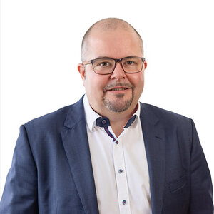 Geschäftsführer Tobias Lang mit Brille und kurzem Kinnbart blickt frontal in die Kamera, lächelt leicht. Trägt dunkelblauen Anzug und weißes Hemd. Neutraler, weißer Hintergrund, Oberkörperporträt, professionelle Studioausleuchtung.