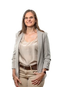 Erwachsene Person mit Brille steht frontal, blickt in die Kamera; trägt hellgraues Sakko, beigefarbenes Seidentop, helle Hose mit braunem Gürtel und Uhr; rechte Hand in Hosentasche; weißer Studiohintergrund.