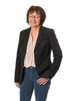 Person mittleren Alters mit kurzer brauner Frisur und Brille steht frontal, trägt schwarzen Blazer, gestreifte Bluse und blaue Jeans; rechte Hand in der Tasche, neutraler weißer Studiohintergrund.