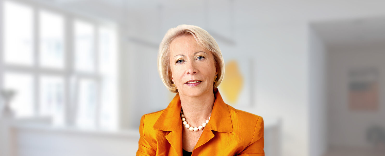 Dr. Annelie Scheuernstuhl mit kurzem blondem Haar – blickt neutral in die Kamera – in hellem, unscharfem Innenraum mit großen Fenstern; trägt orangenes Sakko, Perlenkette und Ohrringe.